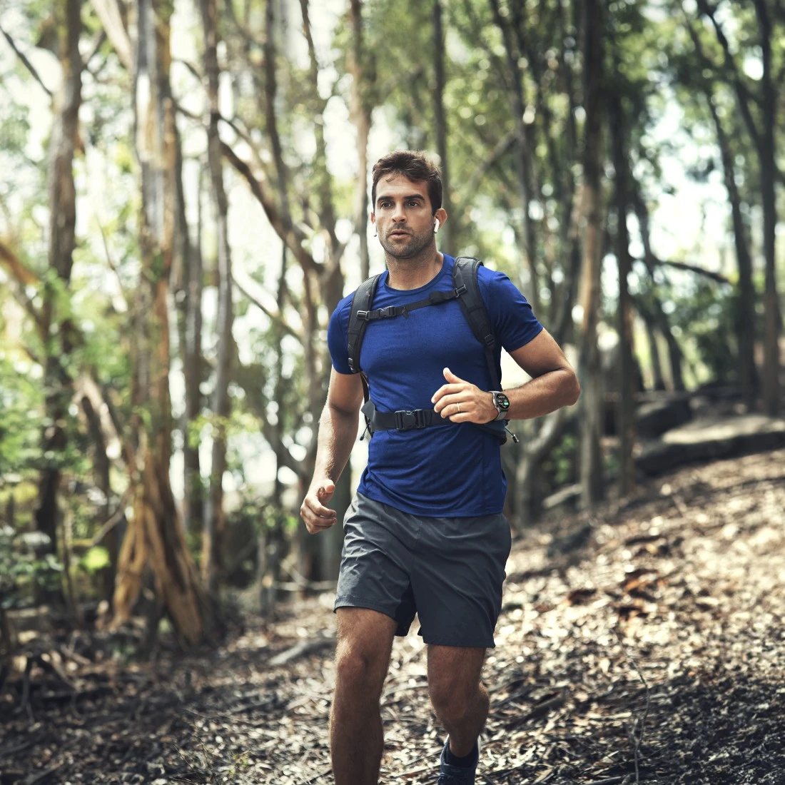 Ein Mann rennt durch einen Wald, trägt ein blaues Hemd, graue Shorts und einen Rucksack. Bäume umgeben ihn, und es ist Tag.