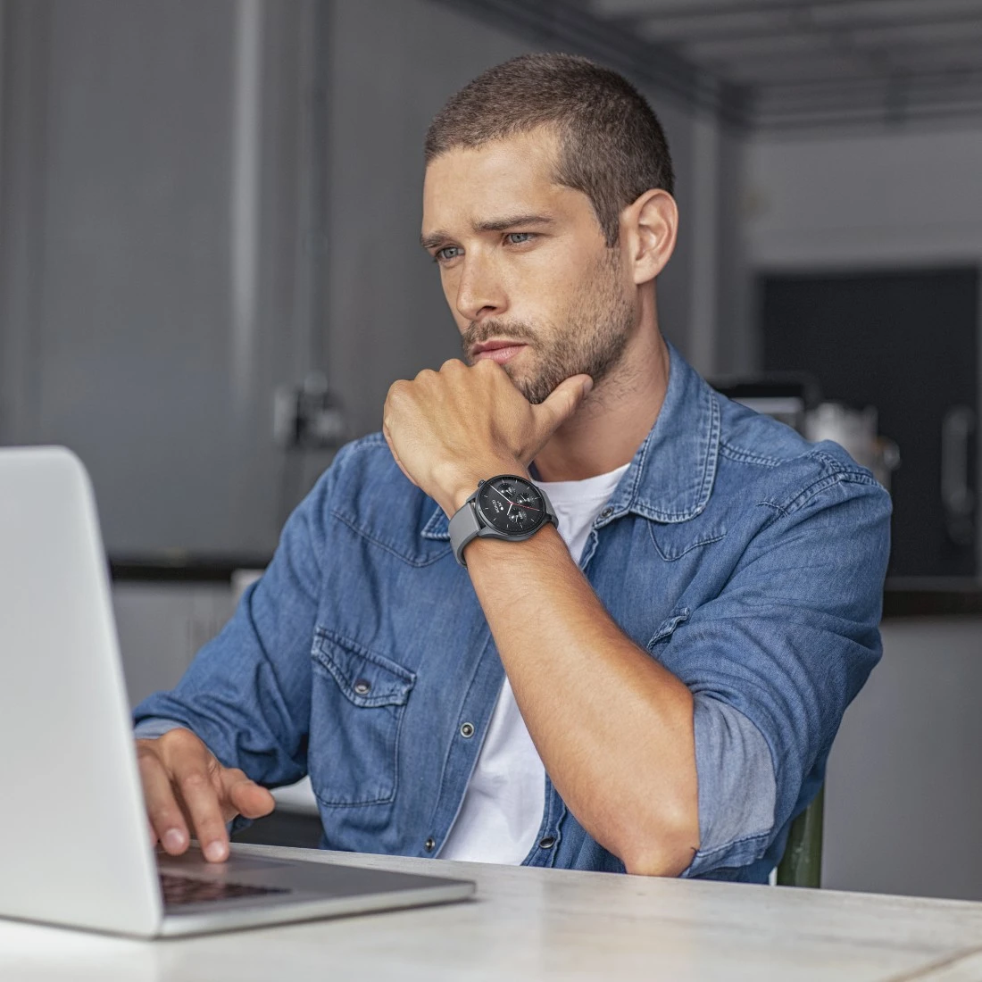 Ein Mann in Jeanshemd betrachtet einen Laptop und stützt sein Kinn auf seine Hand. Er trägt eine Uhr.