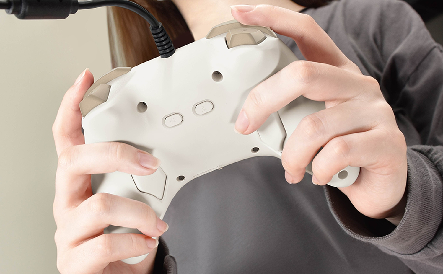 Une personne tient une manette de jeu blanche et beige connectée avec un câble noir. Mains visibles. Chemise grise en arrière-plan.