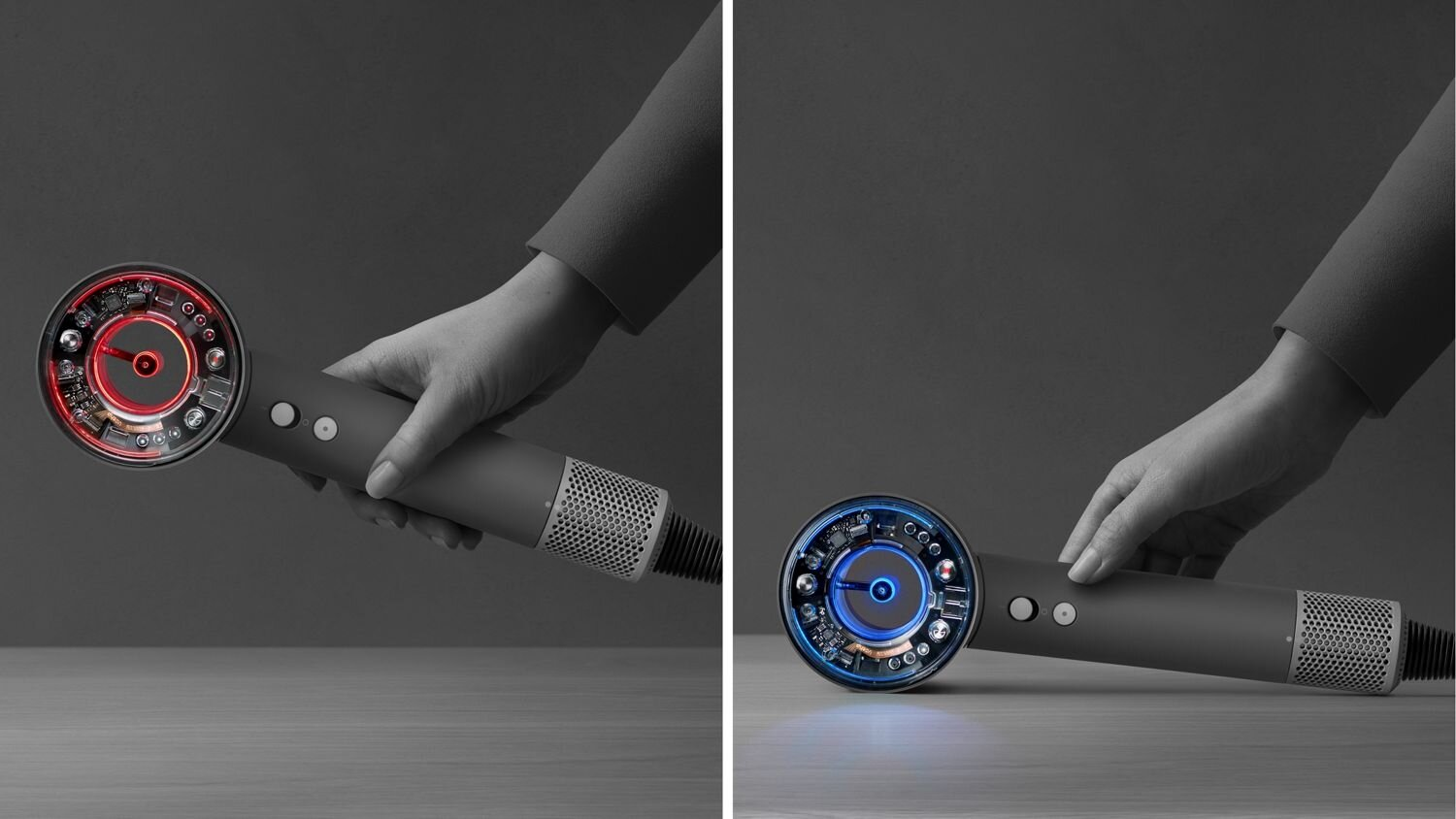 Szara suszarka do włosów z czerwonymi i niebieskimi światłami trzymana jest przez dłoń. Tło jest szare, a suszarka znajduje się na drewnianej powierzchni.