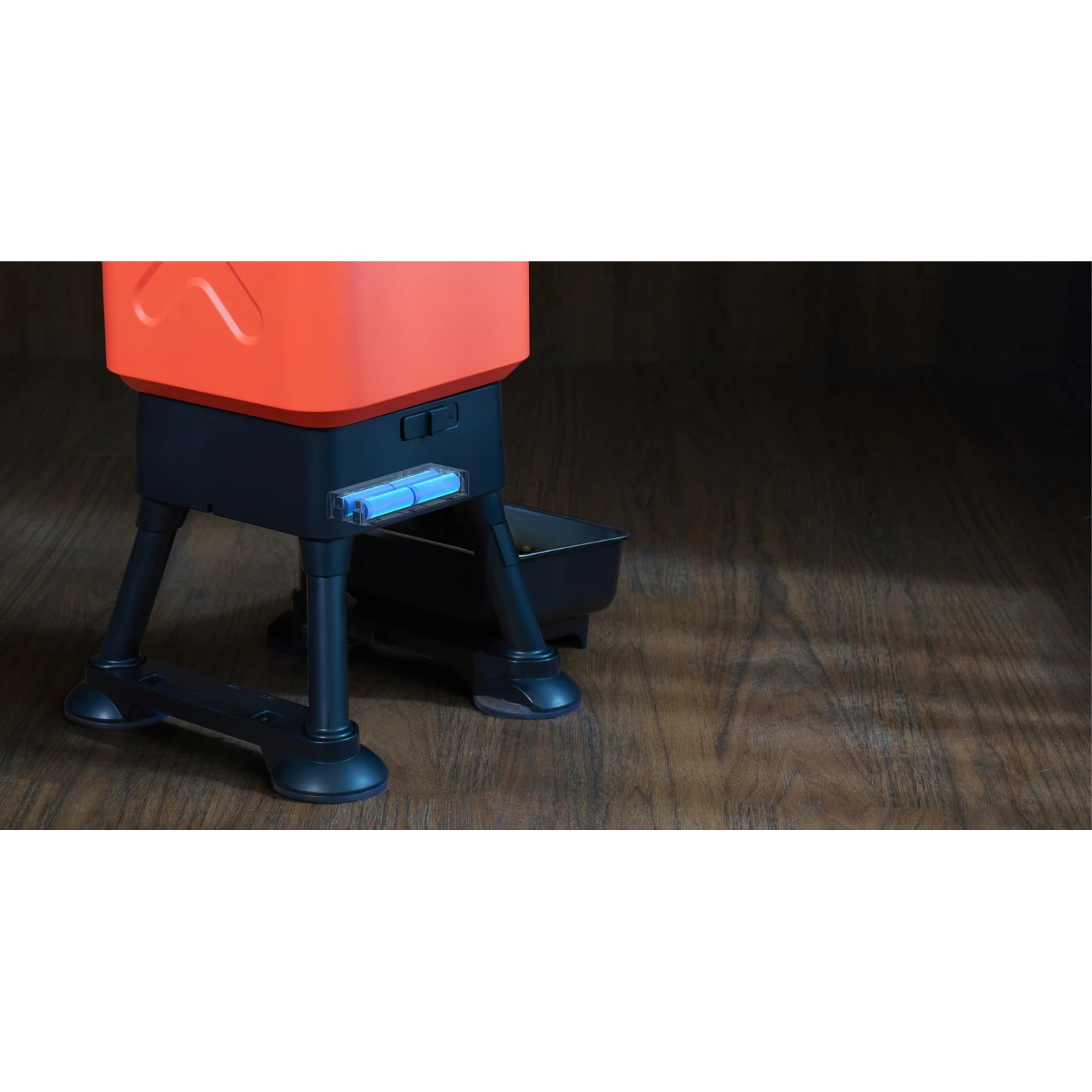 Un alimentatore per animali domestici arancione e nero su un pavimento di legno. L'alimentatore ha un vassoio ed è sostenuto da gambe.