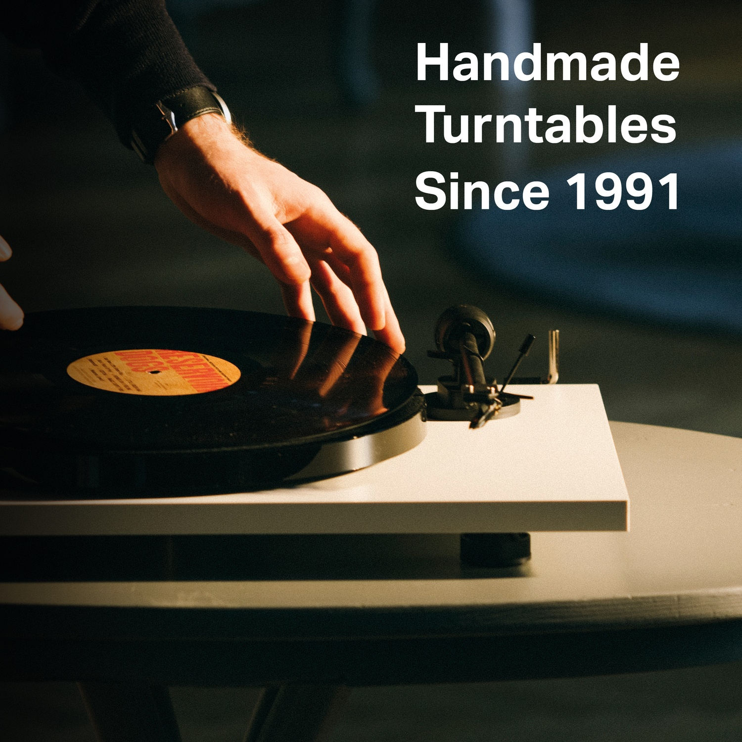 Una mano posiziona un disco in vinile su un giradischi. Il giradischi è su un tavolo rotondo. Testo: Handmade Turntables Since 1991.
