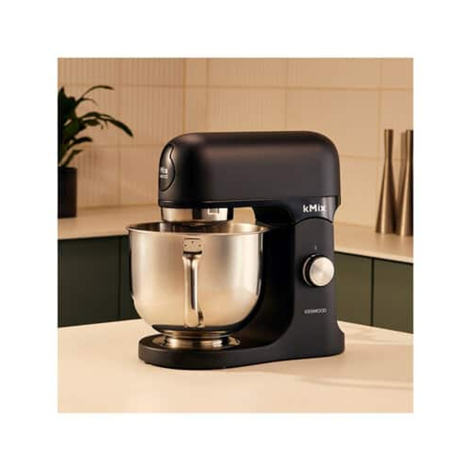Un mixer a immersione Kmix nero con una ciotola argentata è posto su una superficie bianca. Lo sfondo mostra un ambiente cucina.