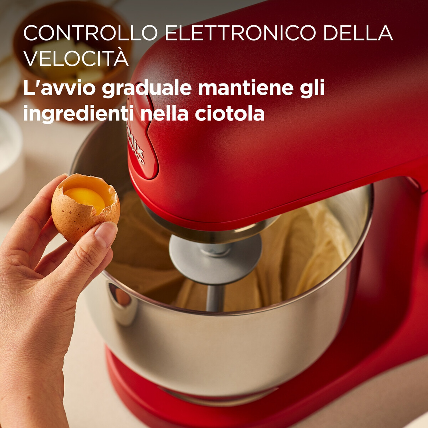 Una mano rompe un uovo sopra una ciotola rossa del mixer. Il mixer è rosso. La ciotola è in acciaio inox e contiene pastella.