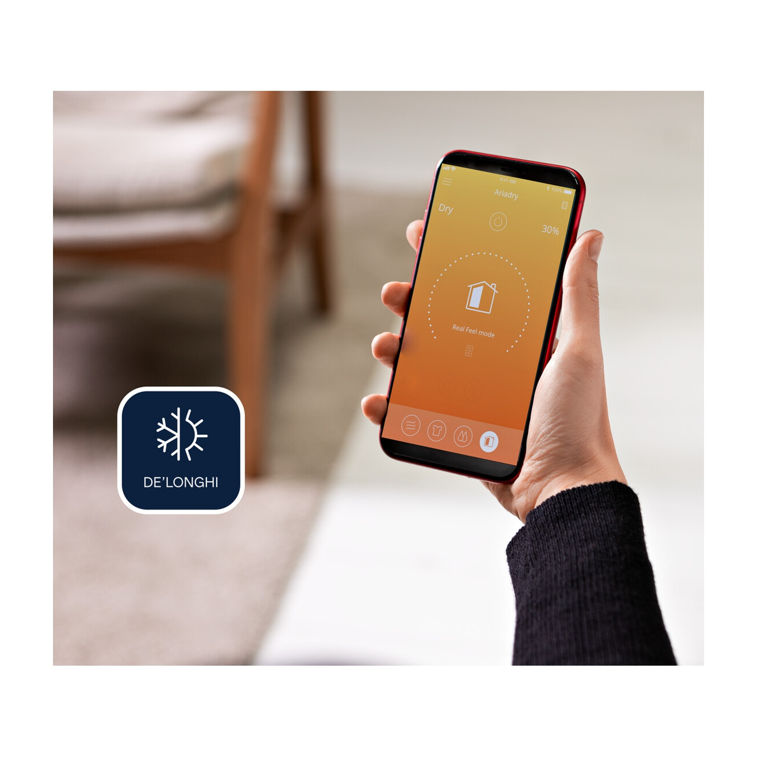 Una mano che tiene un telefono rosso che mostra un'app per deumidificatore. Il logo De'Longhi è in basso a sinistra.
