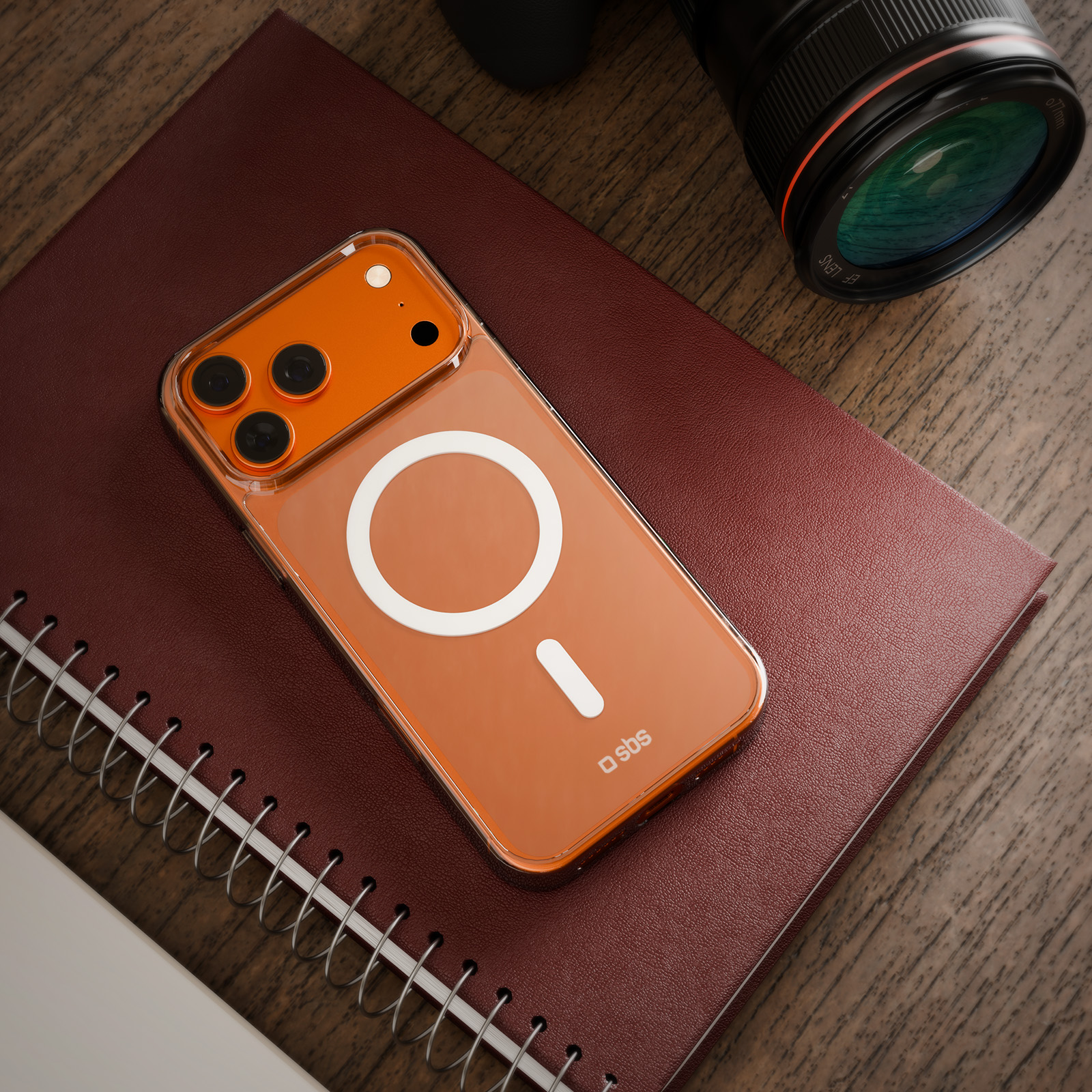 Pomarańczowy iPhone z przezroczystym etui, leżący na bordowym notatniku ze spiralą, obok obiektywu na drewnianej powierzchni.