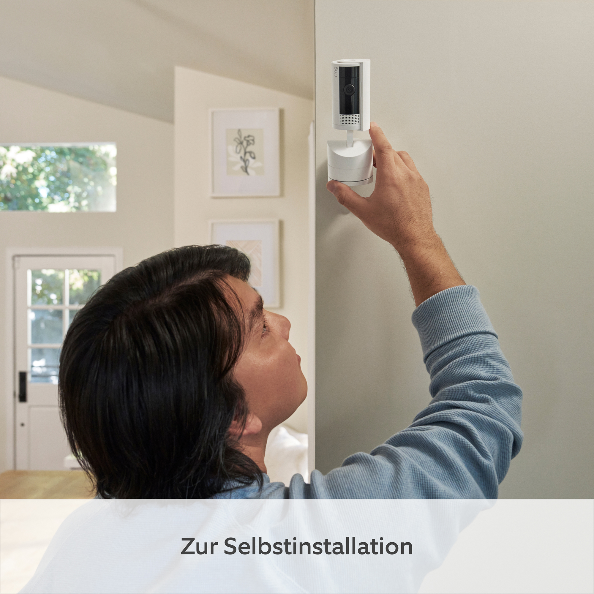 Ein Mann installiert eine weiße Überwachungskamera an einer Wand. Der Mann hat dunkles Haar und trägt ein blaues Hemd.
