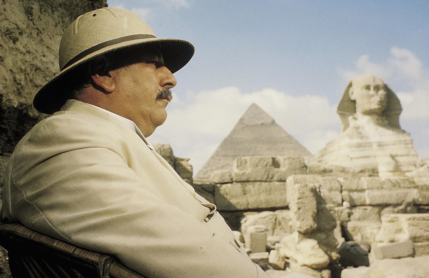 Ein Mann mit Safarihut sitzt im Freien und blickt auf die Sphinx, die Pyramide und den Himmel. Er trägt einen hellen Anzug.