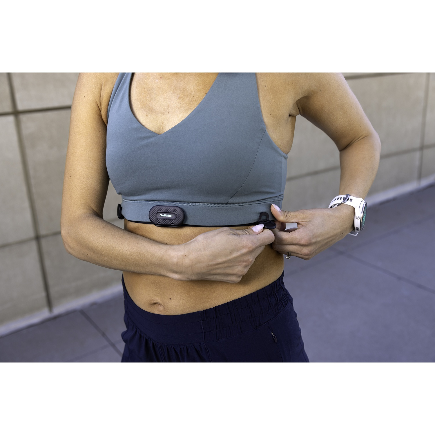 Donna che regola il cardiofrequenzimetro intorno al petto. Indossa reggiseno sportivo, pantaloncini e orologio. Sfondo grigio strutturato.