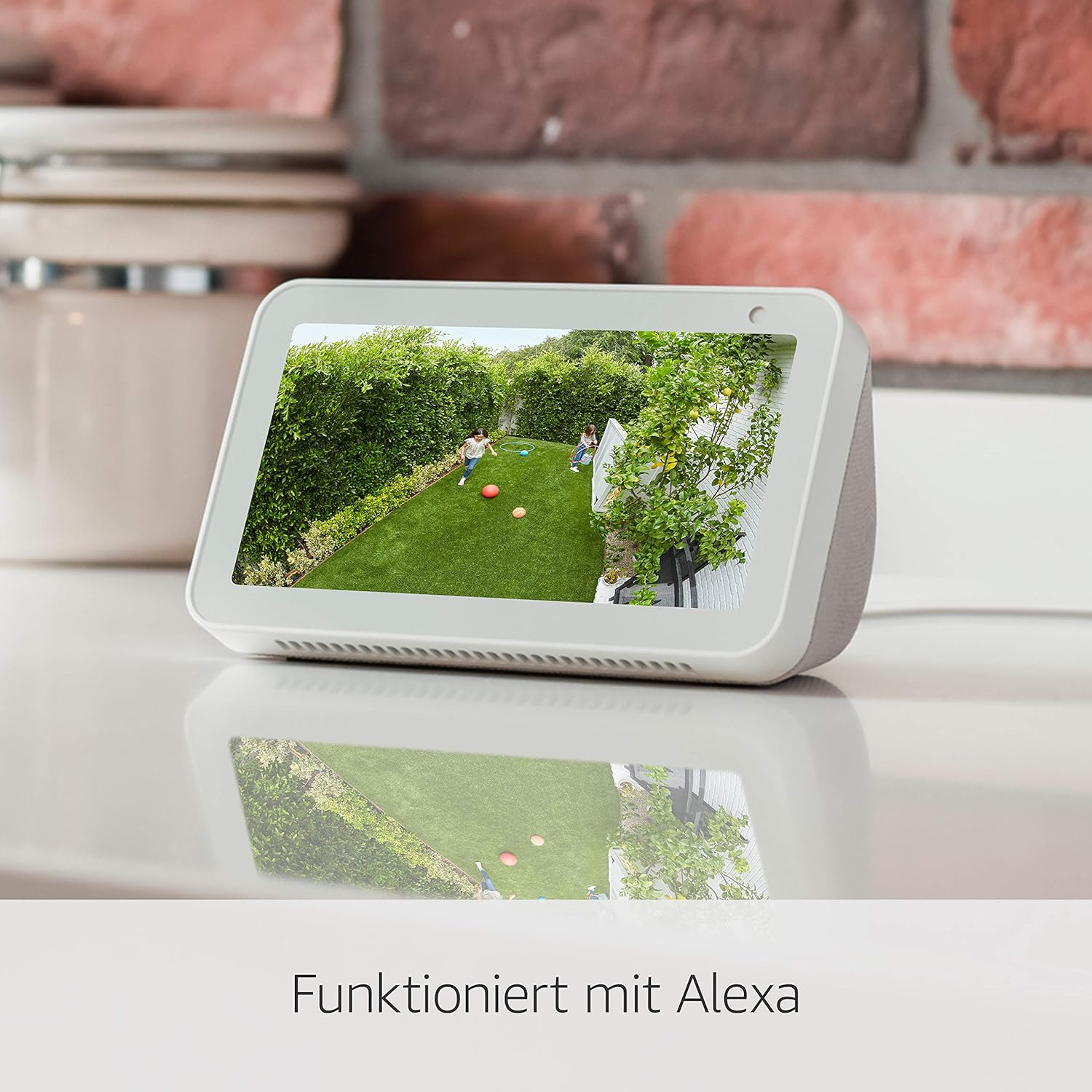 Ein Smart Display zeigt Kinder, die in einem Garten spielen. Das Display steht auf einer weißen Oberfläche mit Ziegelmauer im Hintergrund.