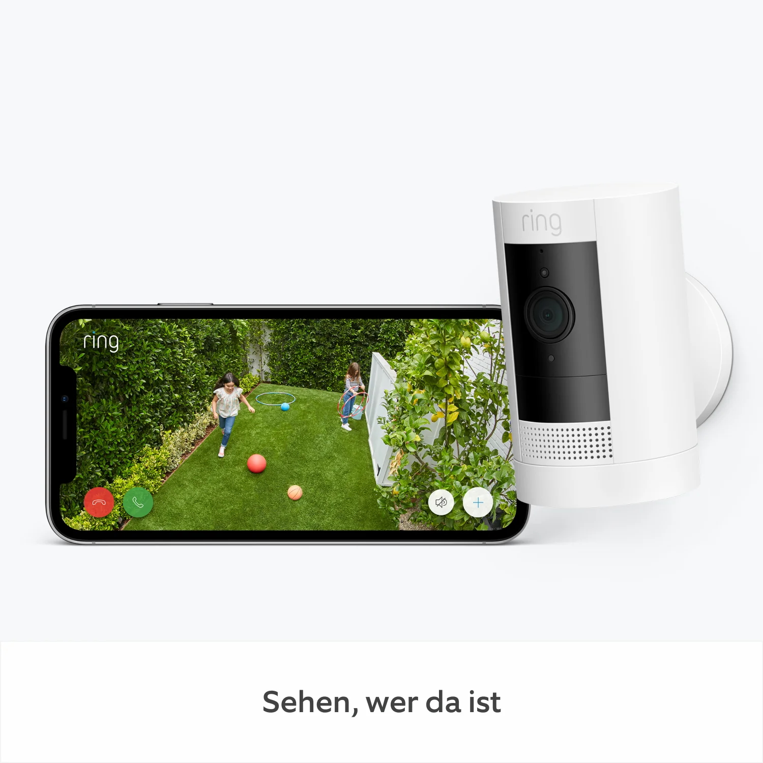Ein Telefon zeigt eine Live-Ansicht der Ring-Kamera von spielenden Kindern auf Gras. Eine weiße Ring-Kamera ist daneben montiert.