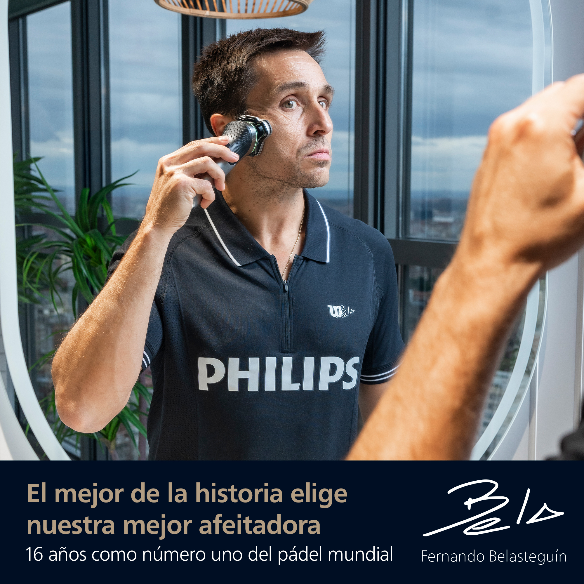 Hombre se afeita con una afeitadora Philips, mirándose en un espejo. Lleva una camisa negra con el logo de Philips. El fondo es una ventana.