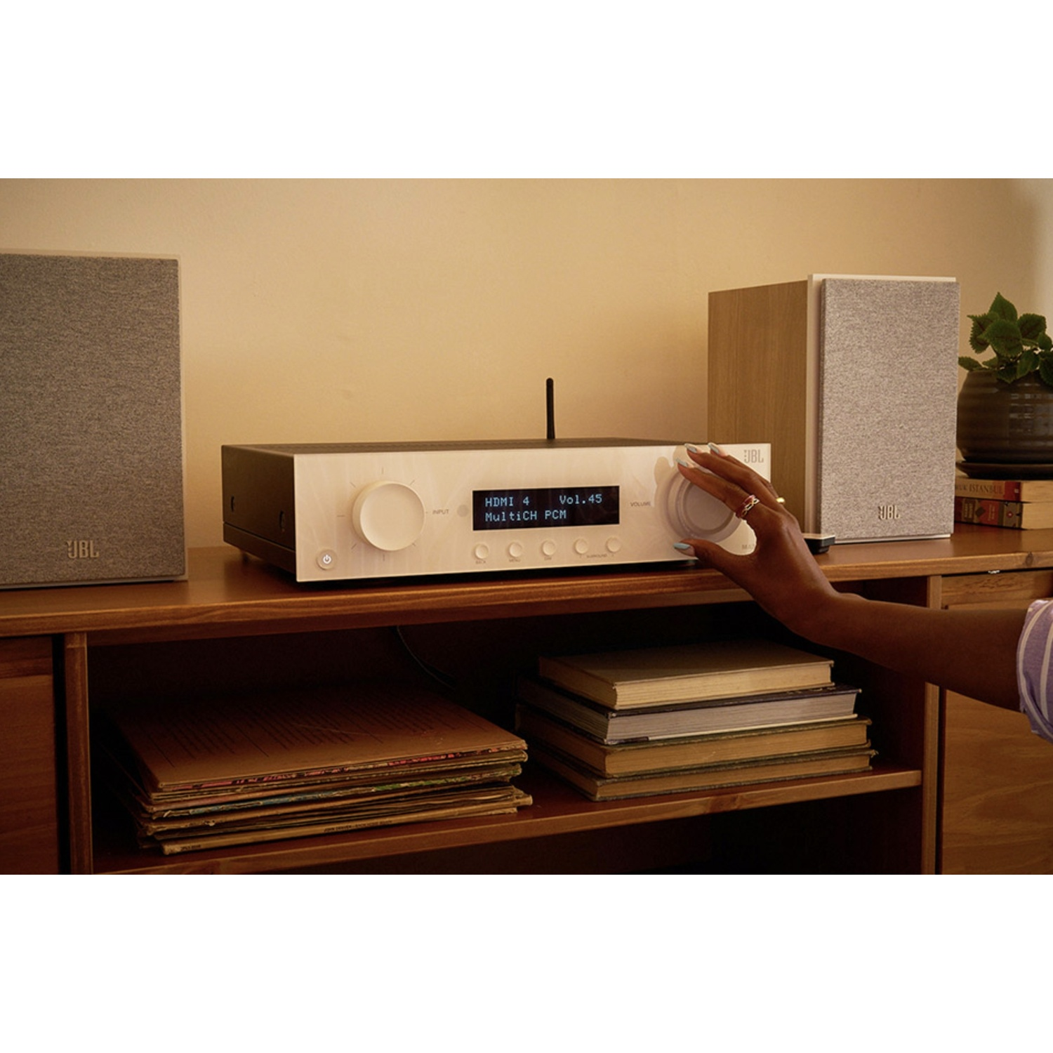 Una persona regola la manopola del volume di uno stereo. Altoparlanti e una pianta sono su una mensola di legno con dischi e libri.