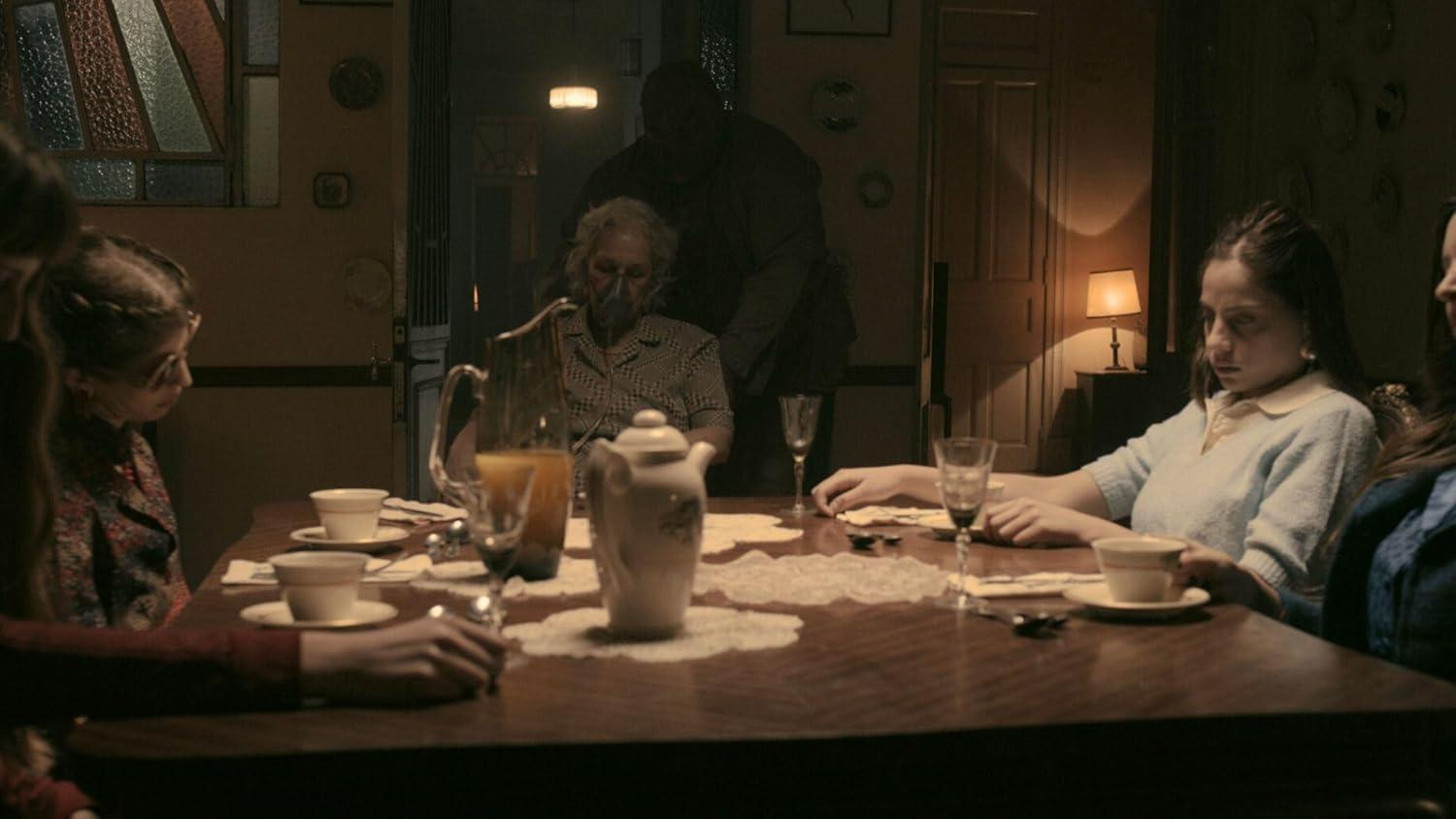 Eine Familie sitzt an einem Tisch. Einem älteren Mann mit Sauerstoffmaske hilft eine große Gestalt. Teetassen, Gläser und eine Lampe sind vorhanden.