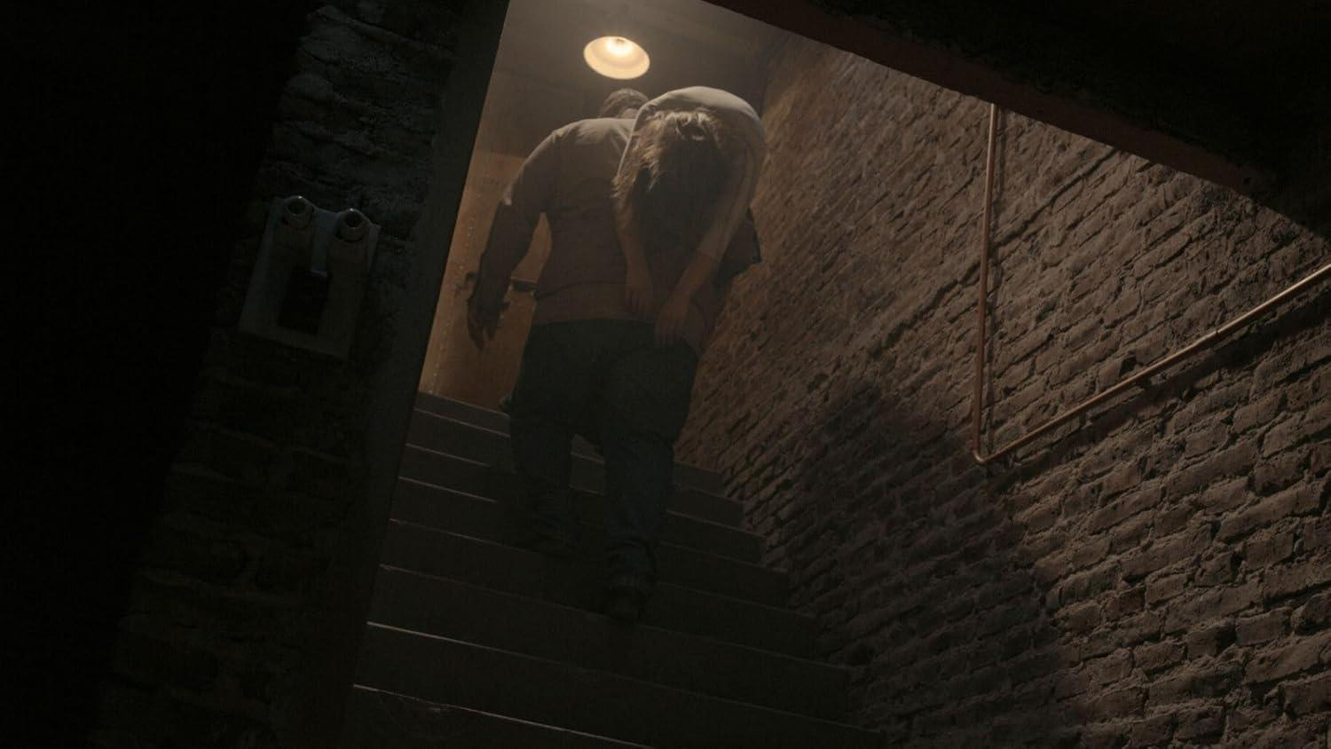 Eine Person trägt eine andere eine schwach beleuchtete Backsteintreppe hinauf. Ein Licht hängt darüber. Die Szene ist dunkel und umschlossen.
