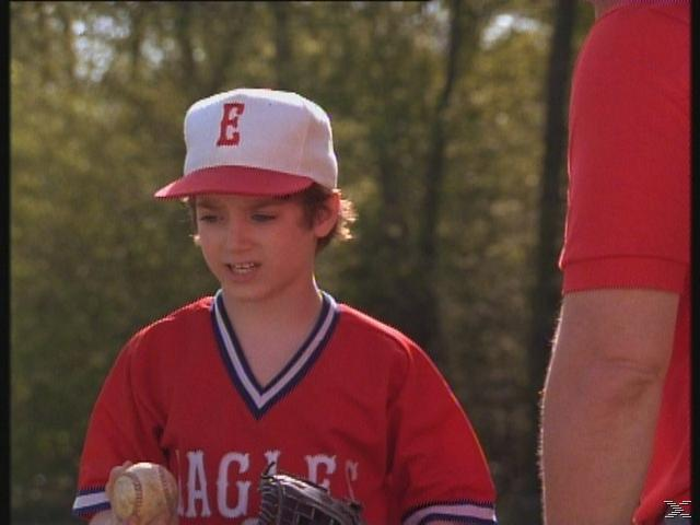 Egy fiatal fiú baseball-egyenruhában labdát tart. 'E' betűs sapkát visel. Egy piros inges férfi jobbra.