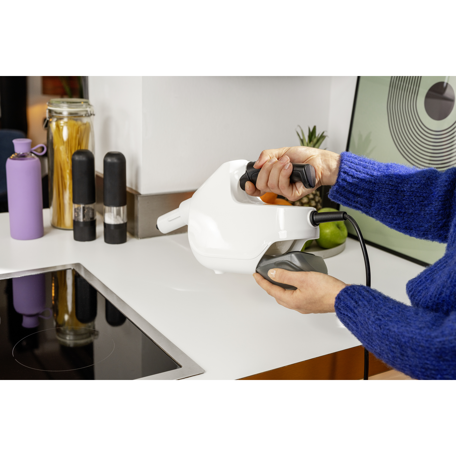 A person in a blue sweater holds a white kitchen appliance. Other objects include a purple bottle, jars, and a green apple.