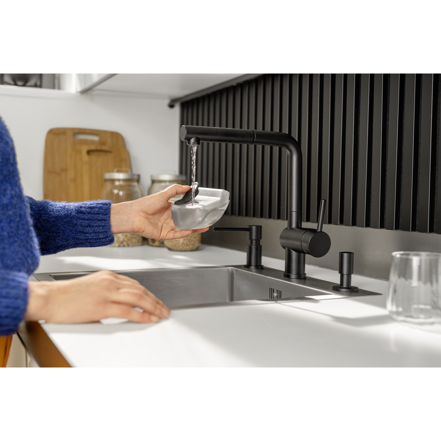 A person fills a container with water from a black faucet. Kitchen sink, black backsplash, and a blue sweater are visible.