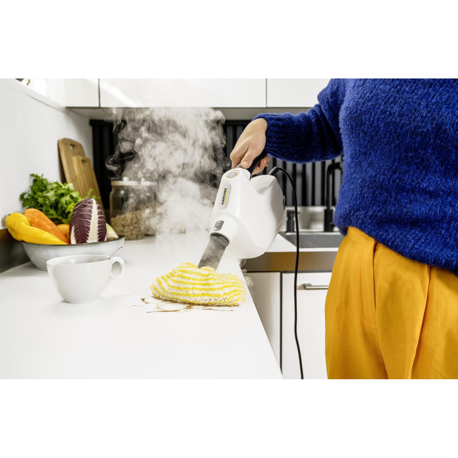A person using a steam cleaner on a white countertop. Vegetables and a cup are on the counter. The person wears a blue sweater and yellow pants.