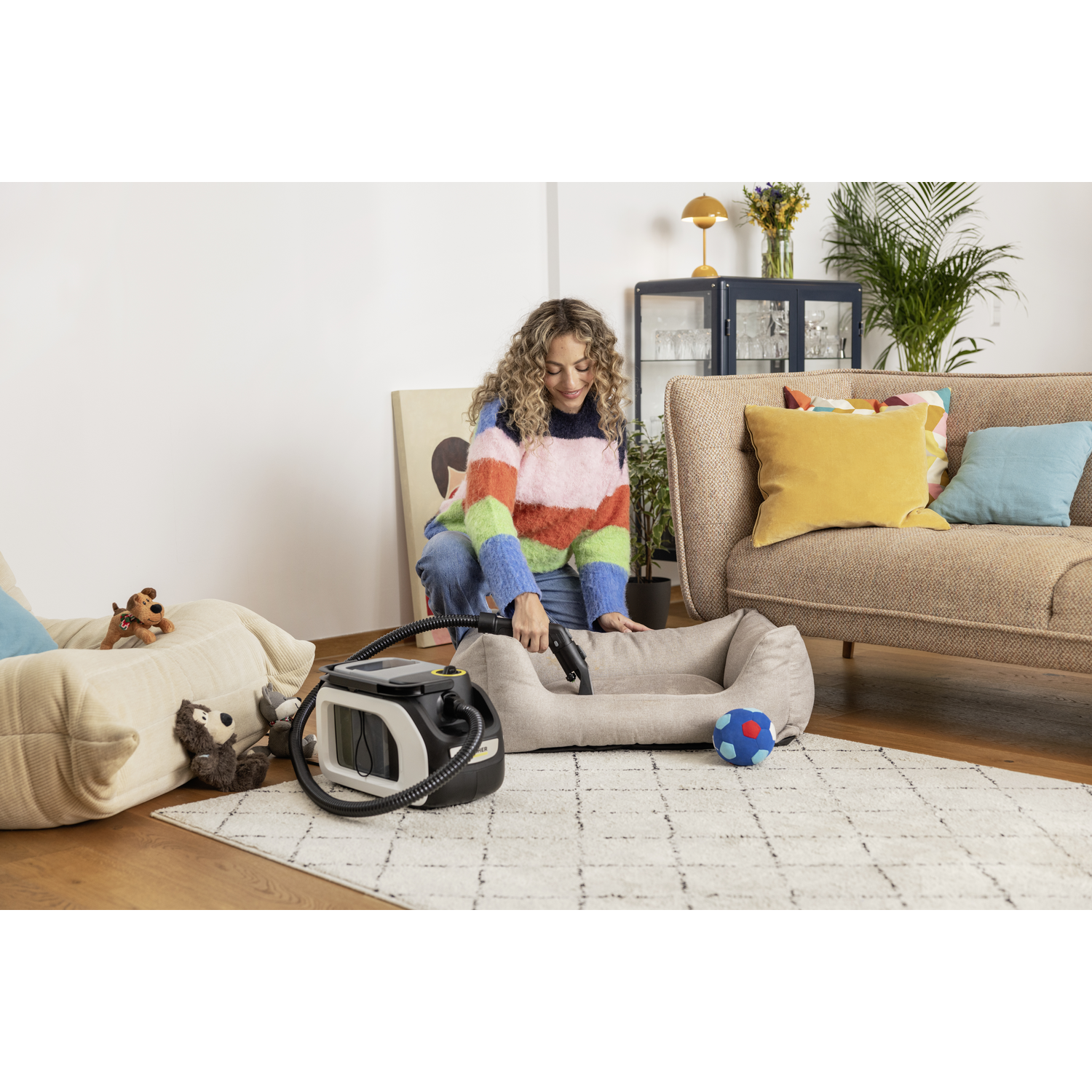 Een vrouw maakt een hondenbed schoon met een stofzuiger. Een bank, speelgoed en een tapijt op de achtergrond.