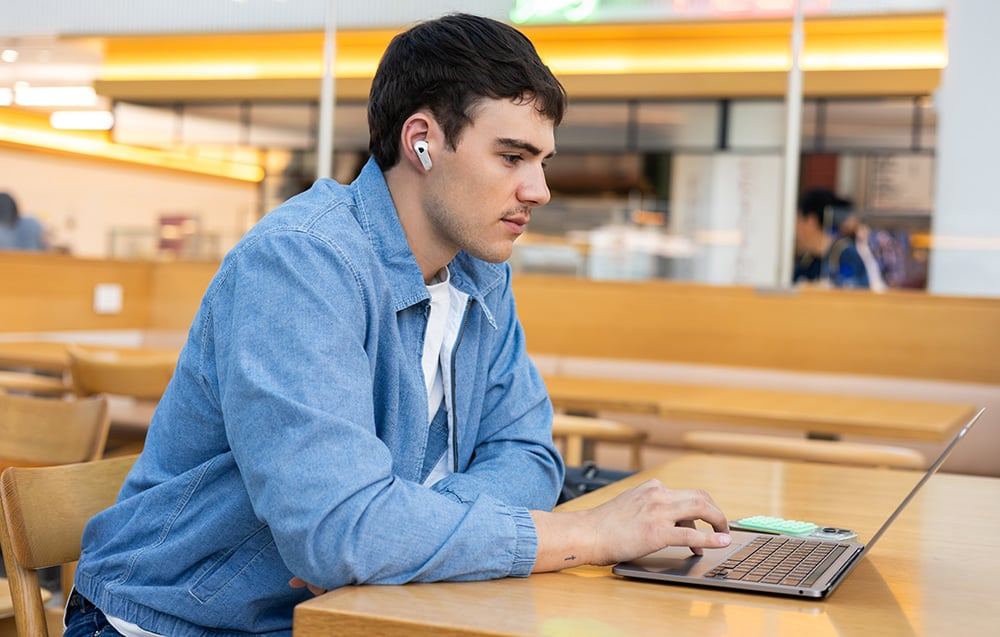 Egy férfi kék farmerdzsekiben laptopot használ egy faasztalnál, fülhallgatóval. Egy kávézóban ül.