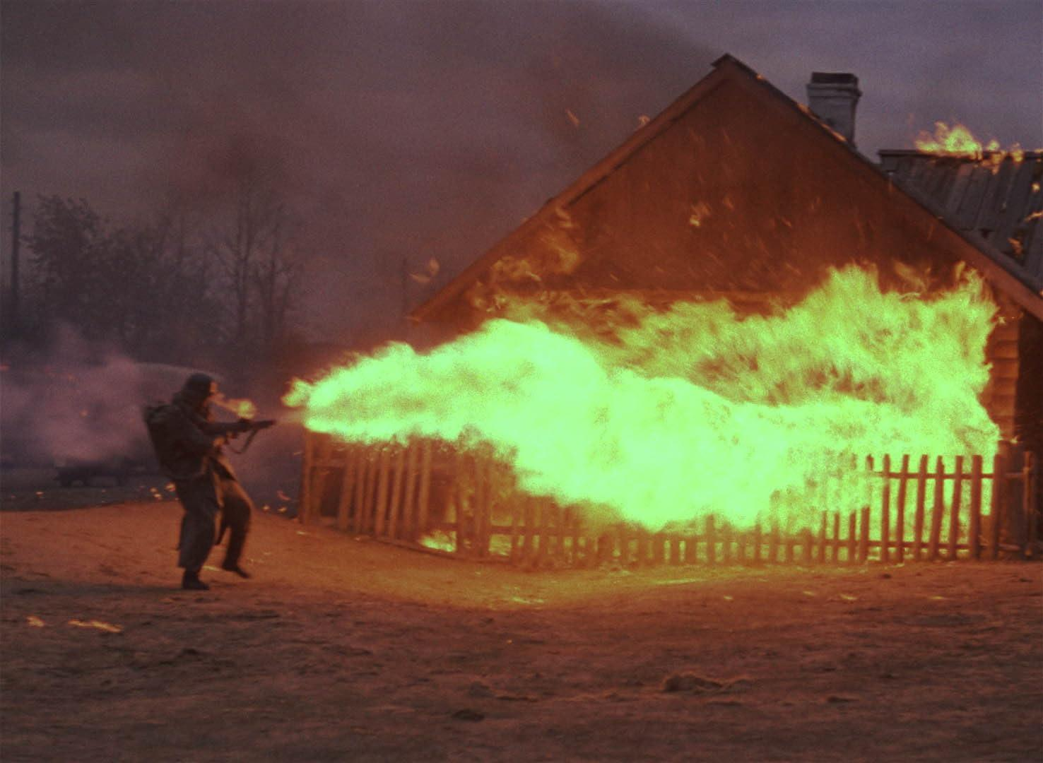 Ein Soldat mit einem Flammenwerfer setzt ein Holzhaus in Brand, Flammen verschlingen die Struktur und den umliegenden Zaun, Rauch erfüllt die Luft.
