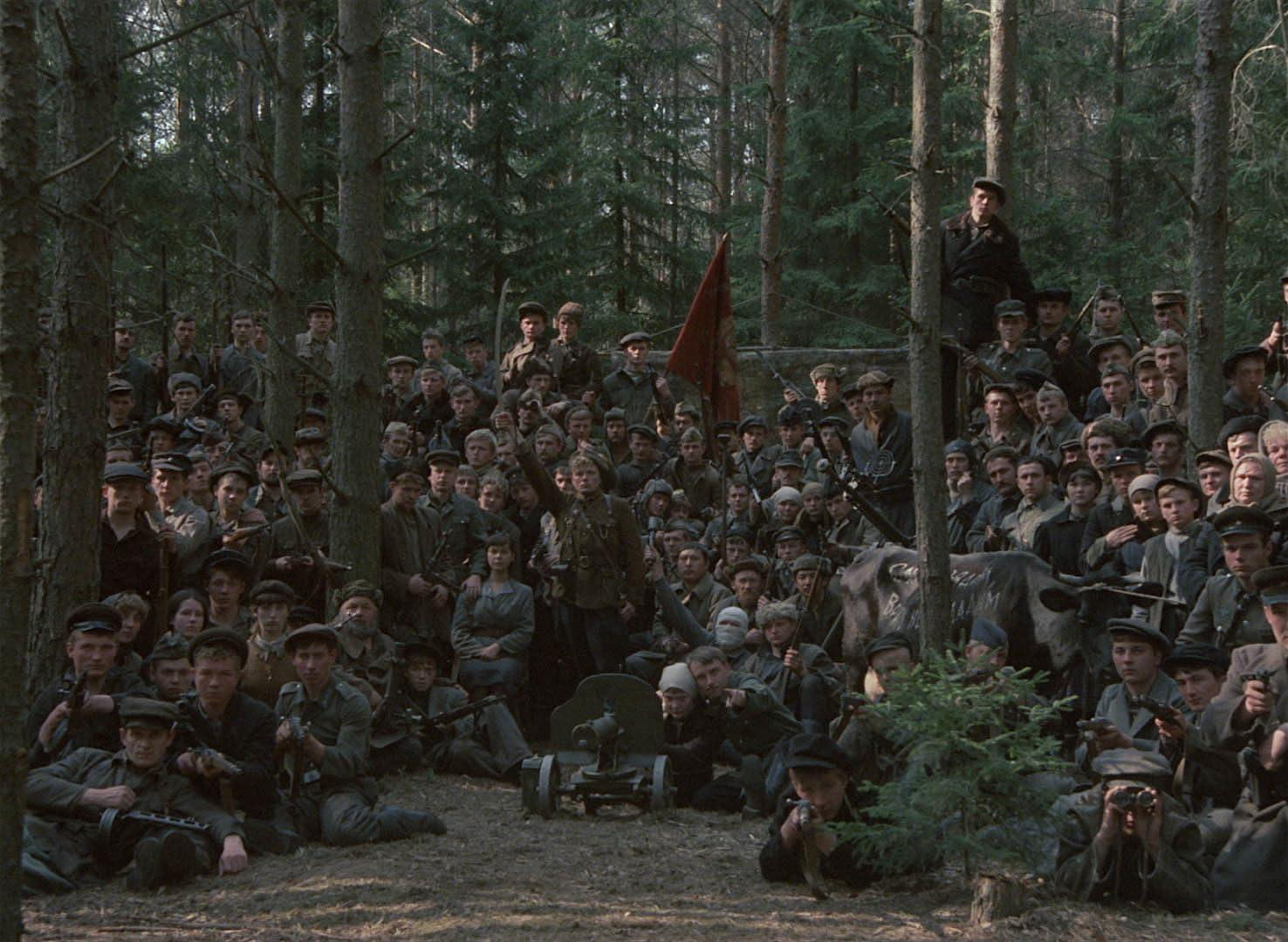 Eine große Gruppe von Männern in Militärkleidung versammelt sich in einem Wald, einige halten Waffen. Eine rote Fahne ist sichtbar.