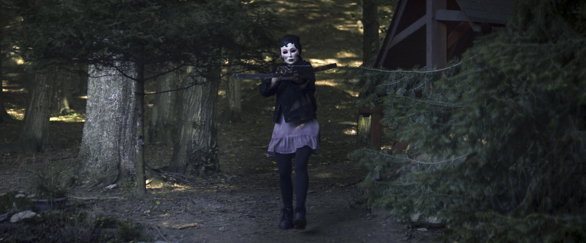 Eine Person mit Maske hält eine Waffe in einem Wald. Dunkle Jacke, rosa Rock, schwarze Leggings und Stiefel. Bäume und Hütte im Hintergrund.