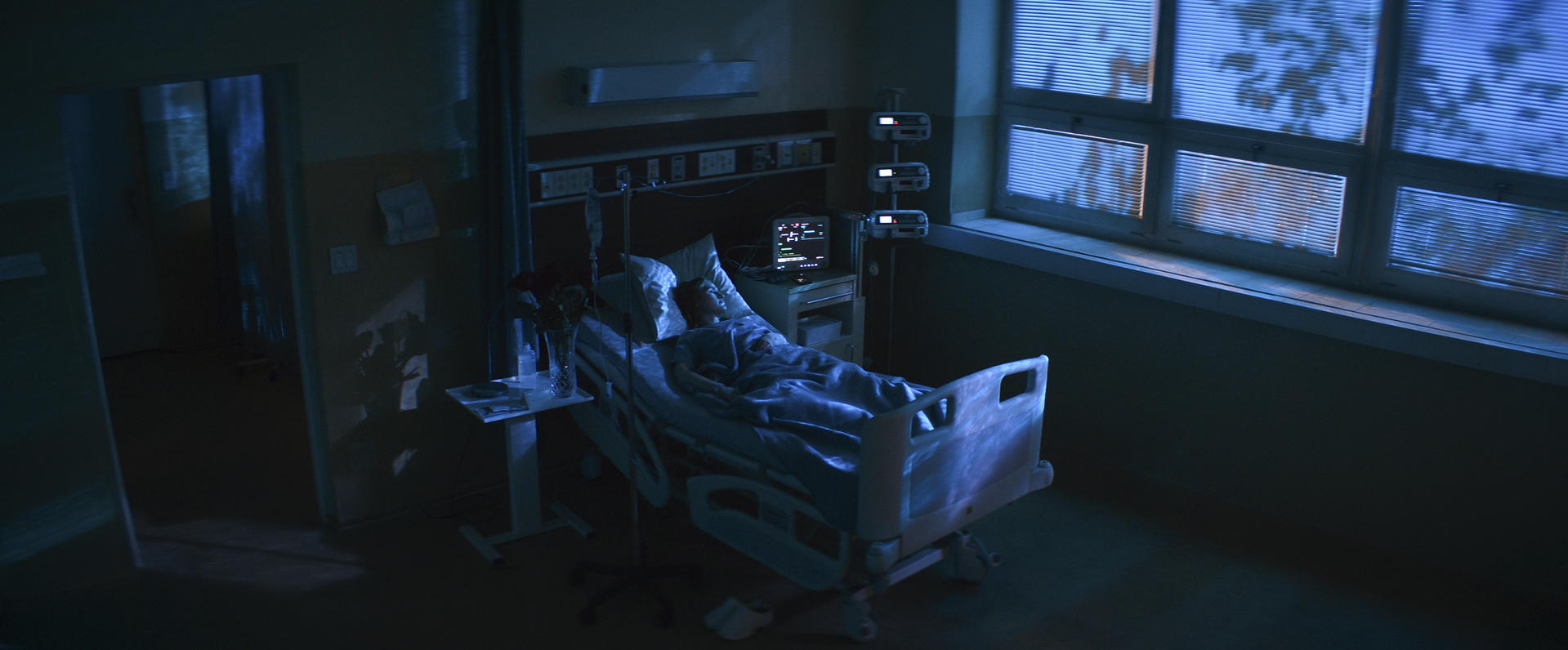 Eine Person liegt in einem Krankenhausbett. Medizinische Geräte umgeben das Bett. Im Hintergrund befindet sich ein Fenster mit Jalousien.