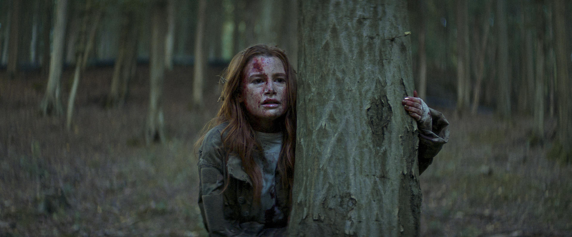 Frau mit Blut im Gesicht versteckt sich hinter einem Baum im Wald. Bäume sind im Hintergrund. Sie trägt eine Jacke.