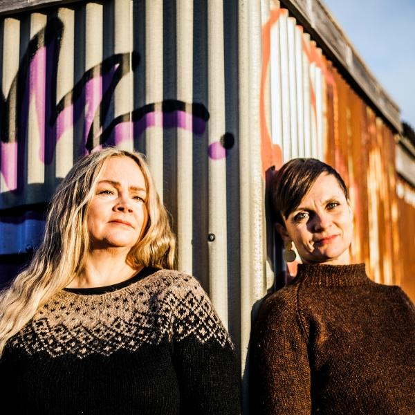 Zwei Frauen stehen an einer Wellblechwand mit Graffiti. Eine trägt einen gemusterten Pullover, die andere einen braunen Pullover. Sonnenlicht scheint.