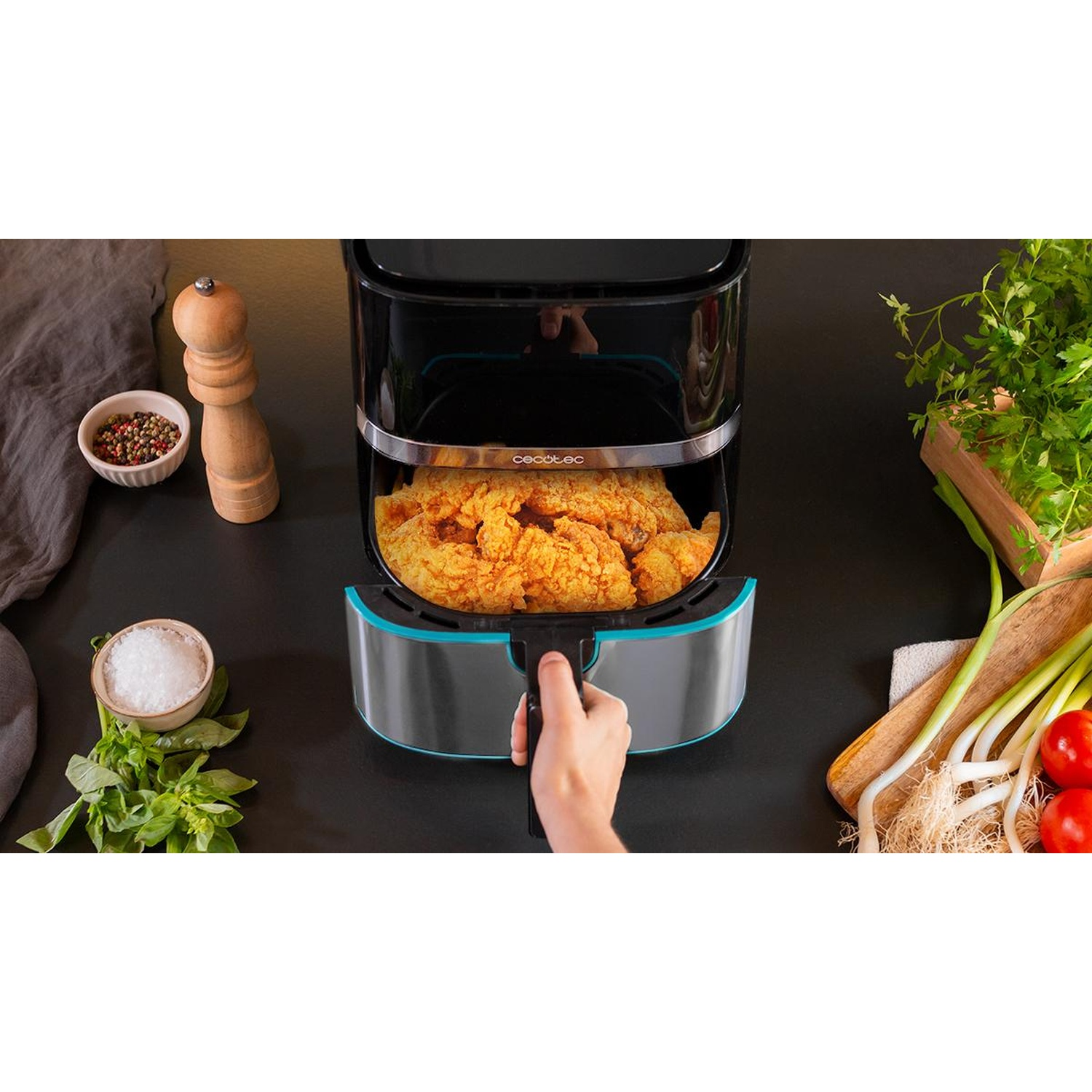 Una friggitrice ad aria con pollo fritto dentro. Vari ingredienti come sale, pepe, erbe aromatiche e verdure sono sul tavolo.