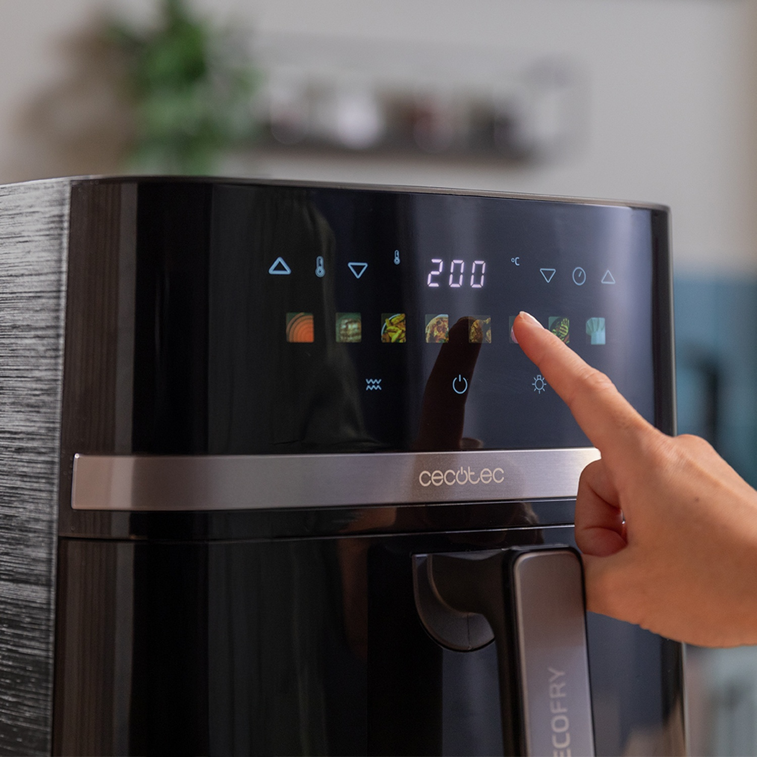 Una mano tocca il pulsante di una friggitrice ad aria con un display digitale che mostra 200 gradi. La friggitrice è nera e argento.