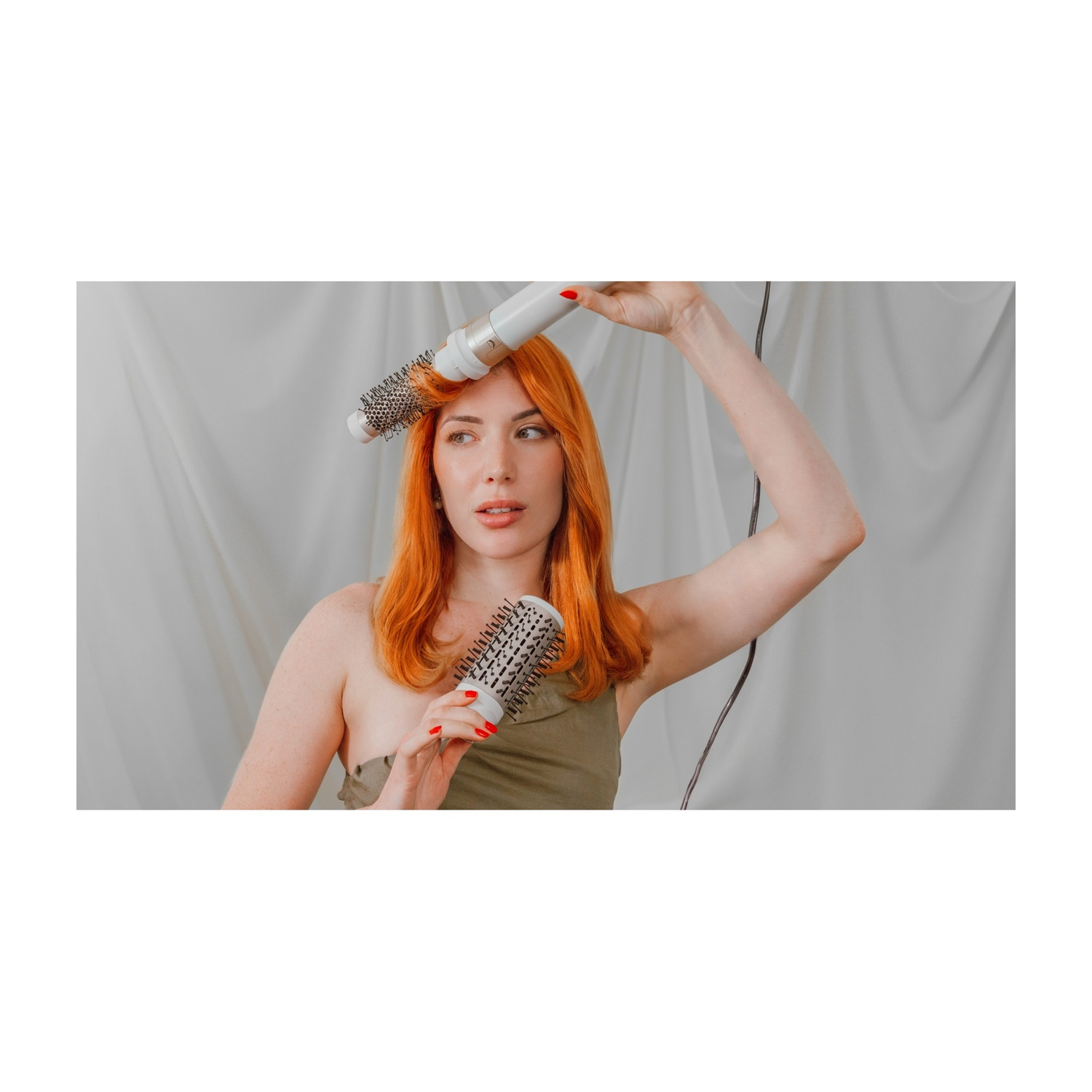 Una donna dai capelli rossi si acconcia i capelli con una spazzola rotonda e un asciugacapelli. Indossa un top verde, su sfondo bianco.