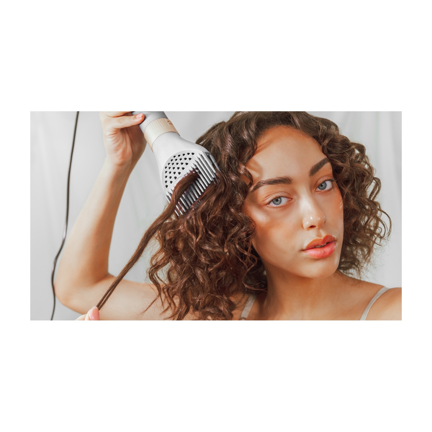 Una donna con i capelli castani ricci si sta acconciando i capelli con un asciugacapelli bianco. Guarda la fotocamera con espressione neutra.
