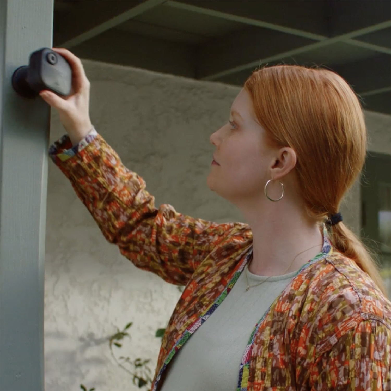 Una donna dai capelli rossi installa una telecamera di sicurezza su un palo blu. Indossa una giacca fantasia e orecchini a cerchio. Sullo sfondo, un muro.