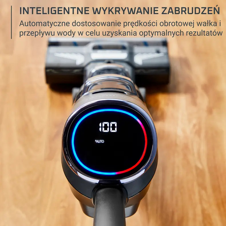 Zbliżenie odkurzacza z cyfrowym wyświetlaczem pokazującym '100' i niebiesko-czerwonym okrągłym wskaźnikiem. Tło to drewniana podłoga.