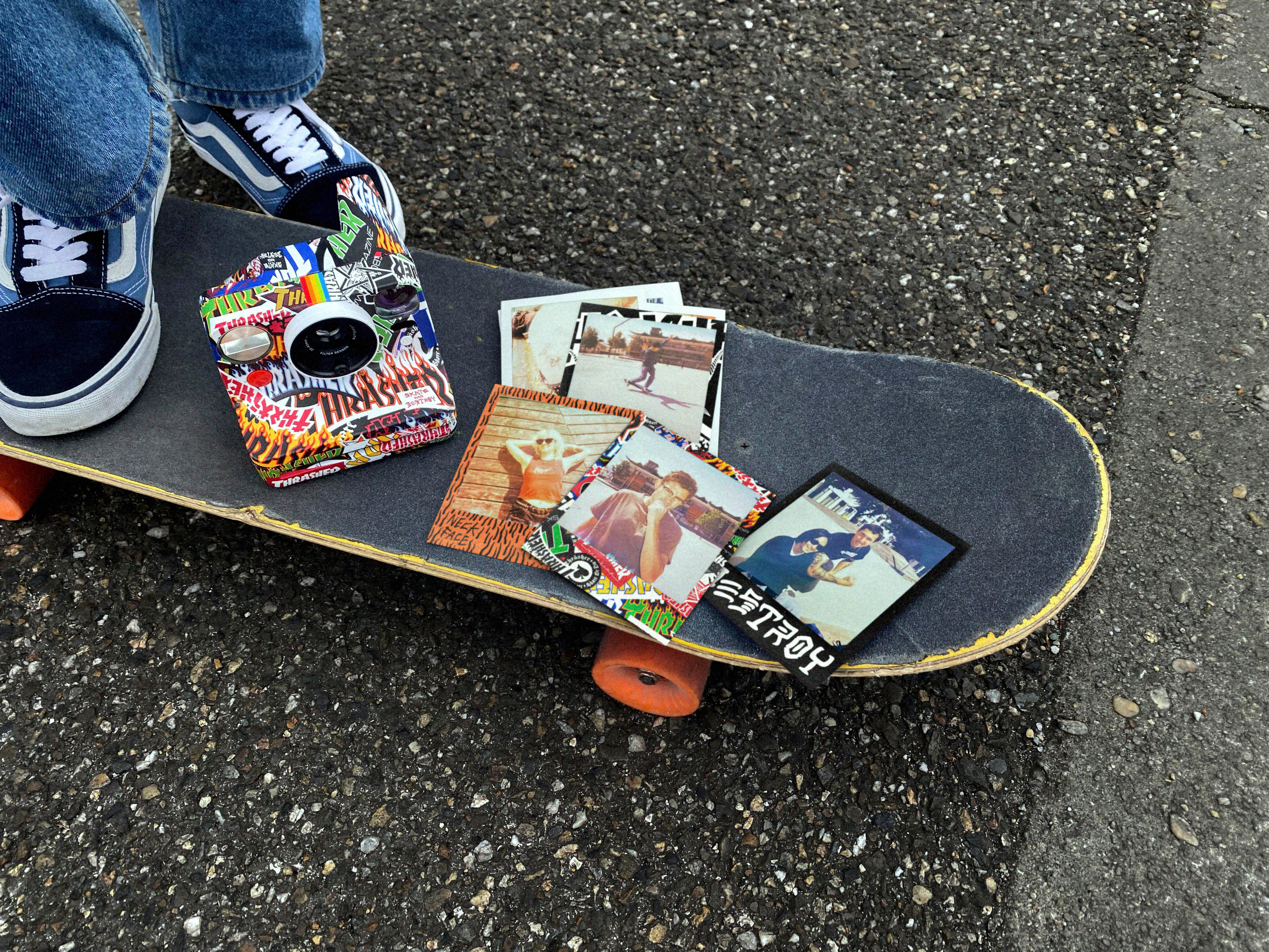 Personenfüße auf einem Skateboard mit Aufklebern, Kamera und Fotos. Der Untergrund ist Asphalt.