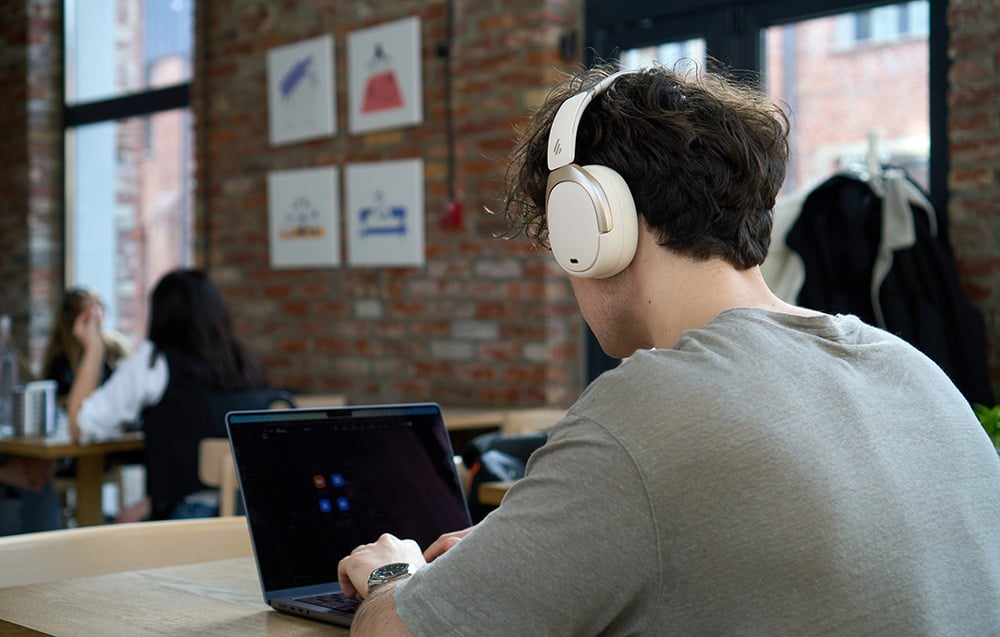 Egy személy fejhallgatóval laptopot használ egy kávézóban. Más emberek és műalkotások a háttérben.