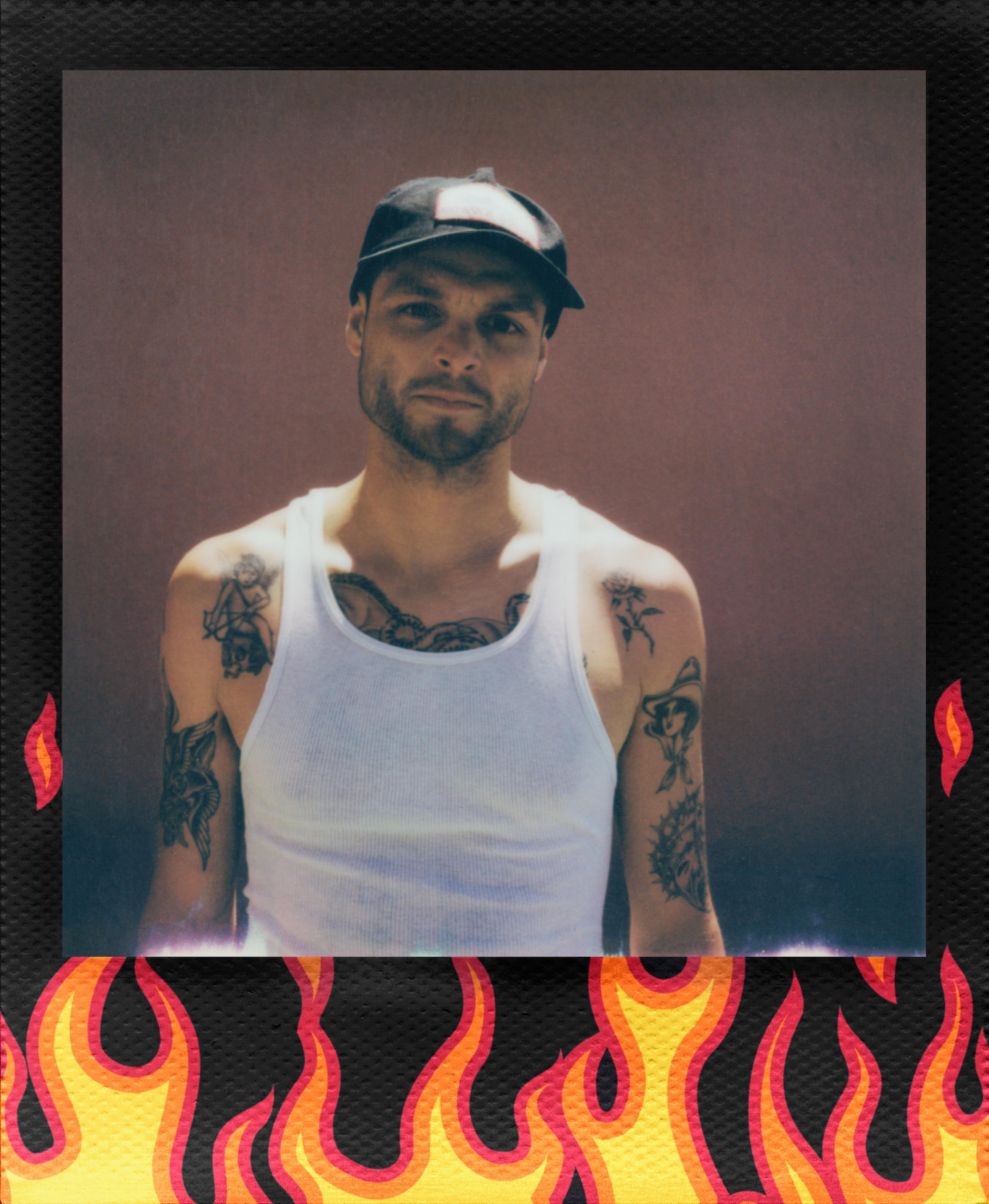 Ein Mann in einem weißen Tanktop und einer Kappe mit Tätowierungen posiert. Der Hintergrund ist braun. Der Rahmen ist schwarz mit Flammen.