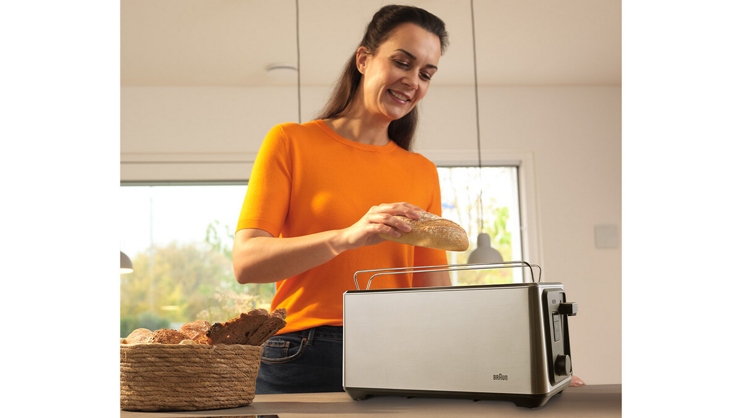 Una mujer con una camisa naranja coloca pan en una tostadora. Una cesta de pan y una ventana están en el fondo.