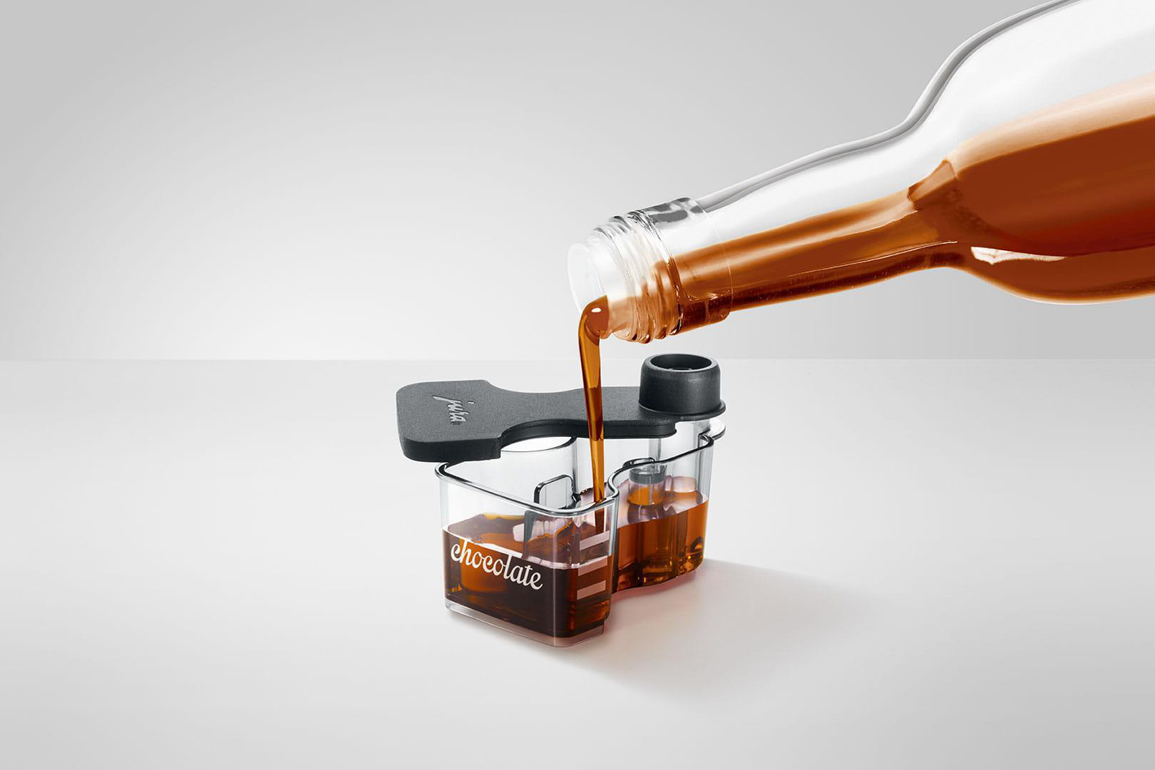 Eine klare Flasche gießt Schokoladensirup in zwei kleine Becher. Die Becher sind mit 'Schokolade' beschriftet und stehen auf einer weißen Oberfläche.
