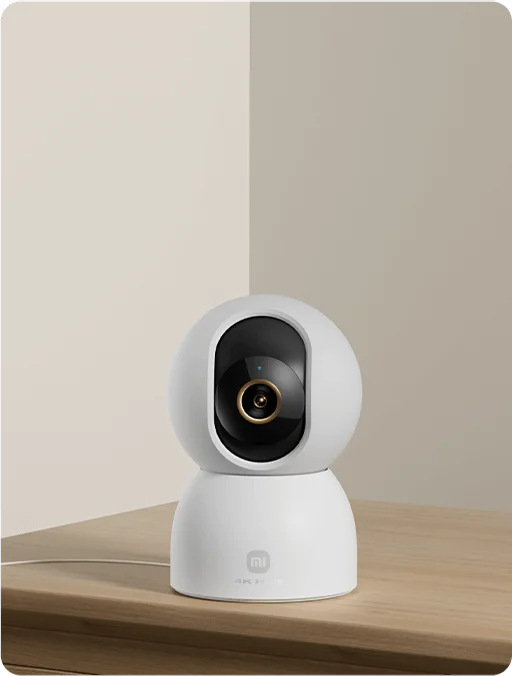 Biała kamera monitoringu na drewnianej powierzchni. Kamera ma czarny obiektyw i złoty pierścień. Tło to jasne ściany.