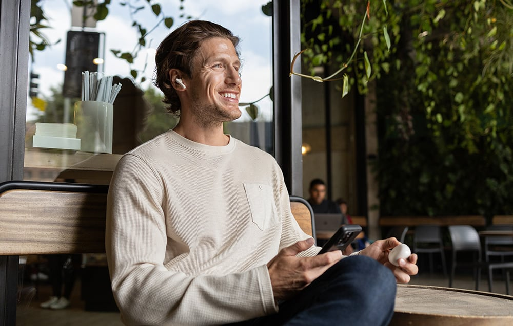 Egy férfi fülhallgatóval mosolyog, telefonnal és egy tojással a kezében egy kávézóban. Zöld lombozat a háttérben. Egy másik személy elmosódott.