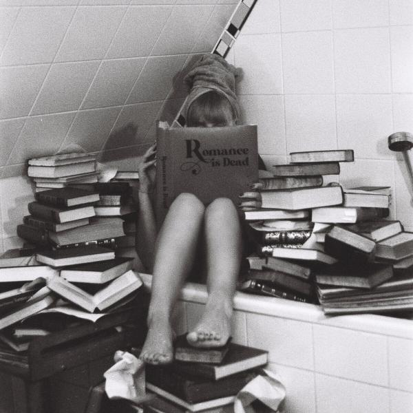 Eine Person sitzt in einer Badewanne, umgeben von Büchern, und liest ein Buch mit dem Titel "Romance is Dead". Die Szene ist schwarz-weiß.
