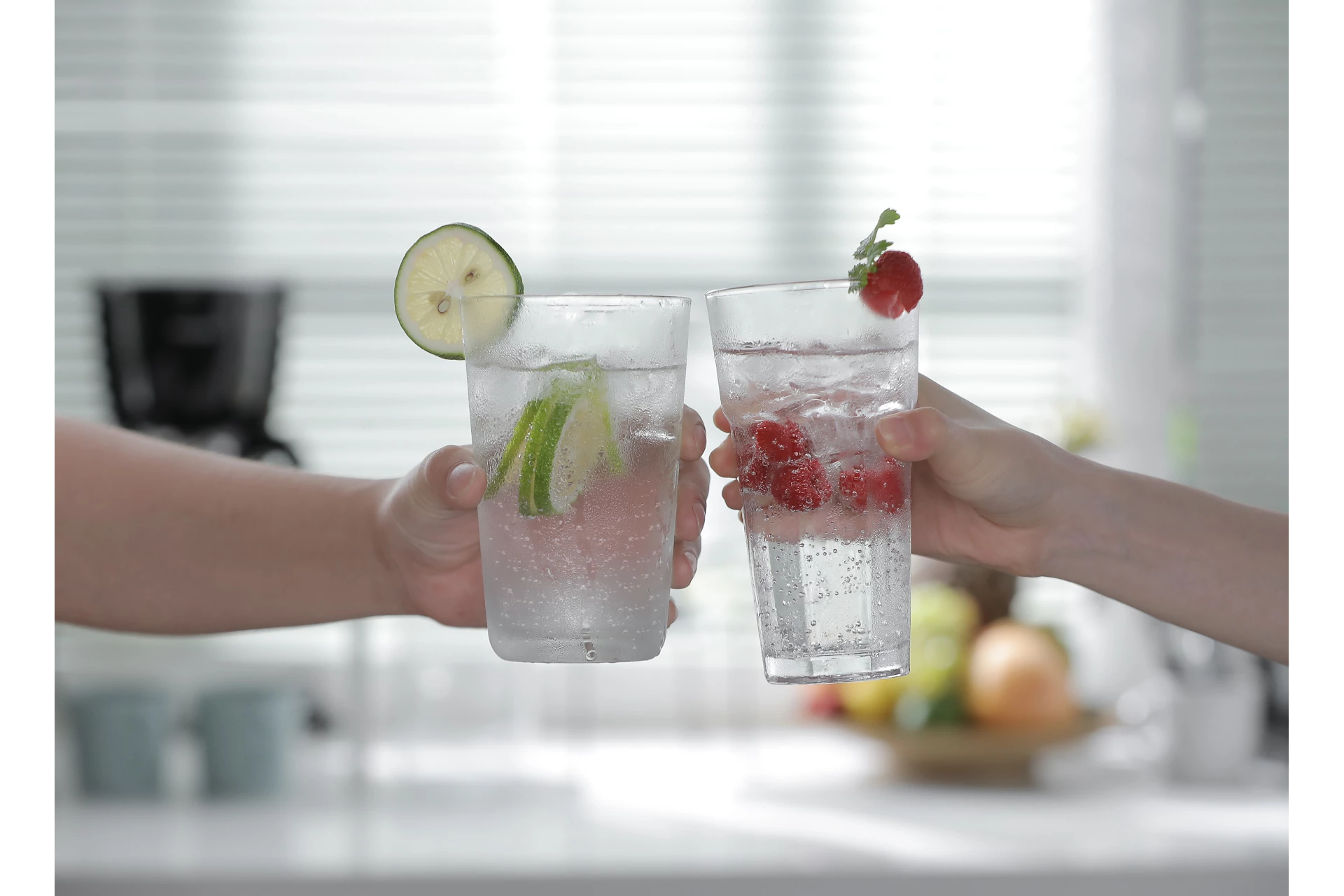 Két kéz tart poharakat lime-mal és málnával. Buborékok láthatók. Konyhai háttér fehér zsaluziákkal.