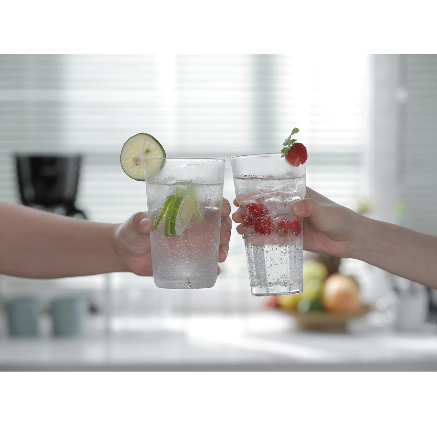Két kéz szénsavas vizet tart, lime-mal és málnával. Konyhai háttér gyümölcsökkel és redőnyökkel.
