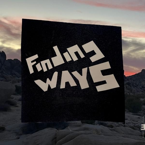 Ein schwarzes Quadrat mit weißem Text 'Finding Ways' schwebt über einer Wüstenlandschaft bei Sonnenuntergang. Berge im Hintergrund.