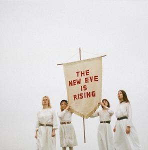 Vier Frauen in weißen Kleidern halten ein Banner mit der Aufschrift 'THE NEW EVE IS RISING' unter bewölktem Himmel.
