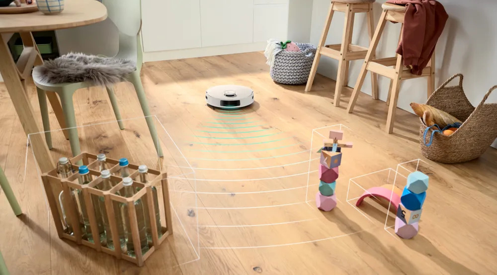 Robot sprzątający porusza się po drewnianej podłodze, omijając przeszkody. Są drewniane stołki, stół i kosz z jedzeniem. Obecne są kolorowe klocki.